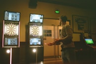 A man is playing darts in a dimly lit room. There are three illuminated electronic dartboards on the wall in front of him, displaying scores and game information. The man is standing to the right, poised to throw a dart. Various posters and a sports jersey are visible on the walls.