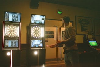 A man is playing darts in a dimly lit room. There are three illuminated electronic dartboards on the wall in front of him, displaying scores and game information. The man is standing to the right, poised to throw a dart. Various posters and a sports jersey are visible on the walls.