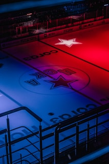 An indoor ice hockey rink is illuminated with dramatic red and blue lighting. The center of the rink showcases a prominent star design, and there is signage along the boards. The foreground features railings, indicating a spectator area.