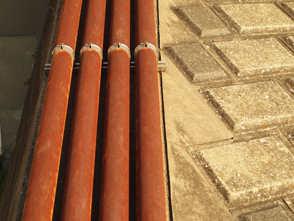 Four horizontal orange pipes are held in place by metal brackets. To the right, a surface comprising square stone tiles arranged in a geometric pattern is visible. The lighting creates warm highlights on the pipes and a textured effect on the tiles.