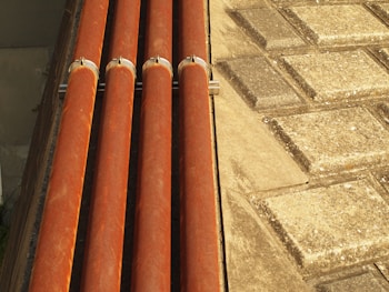 Four horizontal orange pipes are held in place by metal brackets. To the right, a surface comprising square stone tiles arranged in a geometric pattern is visible. The lighting creates warm highlights on the pipes and a textured effect on the tiles.