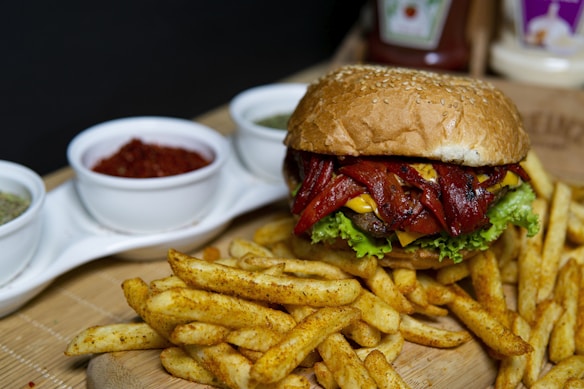 A large cheeseburger with various toppings, including lettuce and grilled red peppers, is placed on a wooden surface. The burger is accompanied by seasoned French fries, and there are three small white bowls containing different sauces or spices in the background.