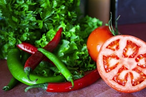 A close-up of fresh vegetables including whole and sliced tomatoes, red and green chili peppers, and leafy greens such as lettuce and parsley. The vegetables are arranged on a wooden surface, displaying vibrant colors and textures.