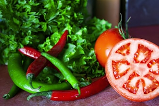 A close-up of fresh vegetables including whole and sliced tomatoes, red and green chili peppers, and leafy greens such as lettuce and parsley. The vegetables are arranged on a wooden surface, displaying vibrant colors and textures.