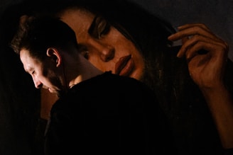 A man is standing in profile, with a contemplative expression, next to a large painting of a woman. The painting features a woman's serene face with closed eyes and an elongated hand placed near her head. The lighting is dim, highlighting the man's face and creating a dramatic contrast with the darker background.