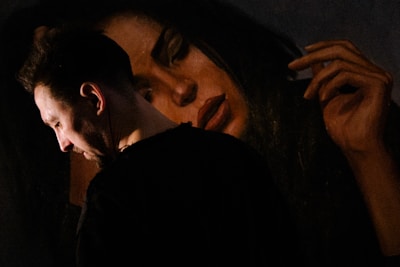 A man is standing in profile, with a contemplative expression, next to a large painting of a woman. The painting features a woman's serene face with closed eyes and an elongated hand placed near her head. The lighting is dim, highlighting the man's face and creating a dramatic contrast with the darker background.