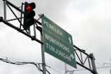 A traffic light displaying a red light is mounted next to a directional road sign. The sign points toward three locations: Peneleh, Wonokromo, and Tunjungan. Overcast skies form the background, and several cables are visible near the sign and light.