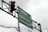 A traffic light displaying a red light is mounted next to a directional road sign. The sign points toward three locations: Peneleh, Wonokromo, and Tunjungan. Overcast skies form the background, and several cables are visible near the sign and light.