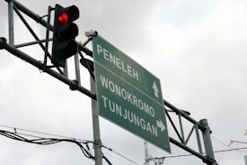 A traffic light displaying a red light is mounted next to a directional road sign. The sign points toward three locations: Peneleh, Wonokromo, and Tunjungan. Overcast skies form the background, and several cables are visible near the sign and light.