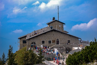 A stone mountain lodge with a gabled roof and a chimney sits atop a rocky hill. Several people are gathered on the terrace, enjoying the scenery. The background is filled with a bright blue sky and soft white clouds. Pine trees are visible in the foreground, adding greenery to the scene.