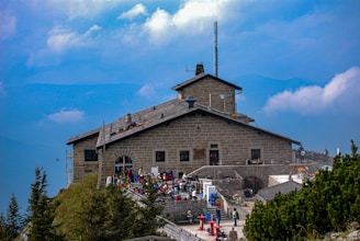 A stone mountain lodge with a gabled roof and a chimney sits atop a rocky hill. Several people are gathered on the terrace, enjoying the scenery. The background is filled with a bright blue sky and soft white clouds. Pine trees are visible in the foreground, adding greenery to the scene.