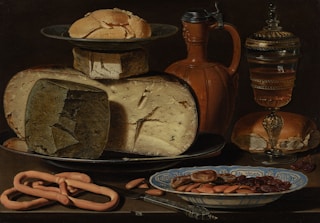 A still life painting featuring various foods including a large wheel of cheese, several smaller cheeses, a mound of butter on a plate, a glass jug, a wineglass with liquid, a loaf of bread, a plate of nuts and dried fruits, and some breadsticks. The items are arranged aesthetically on a table with rich, earthy tones.