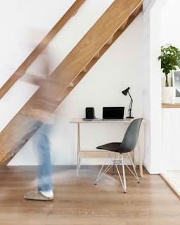 A modern and minimalist home office setup featuring a small wooden desk with a black lamp, a notebook, and a laptop. The desk is positioned against a white wall under a wooden staircase. A black chair with metallic legs is placed next to the desk. A person, slightly blurred due to motion, walks past the setup. There is a potted plant on a shelf in the background.