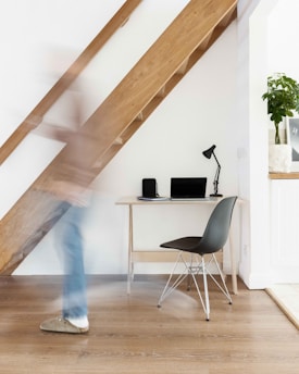 A modern and minimalist home office setup featuring a small wooden desk with a black lamp, a notebook, and a laptop. The desk is positioned against a white wall under a wooden staircase. A black chair with metallic legs is placed next to the desk. A person, slightly blurred due to motion, walks past the setup. There is a potted plant on a shelf in the background.