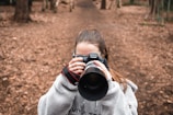 A person in a cozy gray fleece jacket is holding a Canon camera, capturing a photograph in a wooded area. The background consists of a dirt pathway surrounded by trees, covered in fallen leaves.