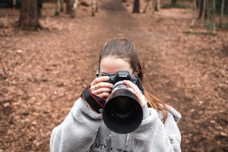 A person in a cozy gray fleece jacket is holding a Canon camera, capturing a photograph in a wooded area. The background consists of a dirt pathway surrounded by trees, covered in fallen leaves.