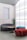 A modern interior space featuring stylish, minimalist furniture including a curved gray sofa, a cylindrical red pouf, and a teal pouf. A round wooden coffee table sits in the center. A large frosted glass wall allows diffused light to fill the room, creating a calm and serene ambiance. A tall green cactus in a pot adds a natural element to the setting.