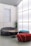 A modern interior space featuring stylish, minimalist furniture including a curved gray sofa, a cylindrical red pouf, and a teal pouf. A round wooden coffee table sits in the center. A large frosted glass wall allows diffused light to fill the room, creating a calm and serene ambiance. A tall green cactus in a pot adds a natural element to the setting.