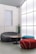 A modern interior space featuring stylish, minimalist furniture including a curved gray sofa, a cylindrical red pouf, and a teal pouf. A round wooden coffee table sits in the center. A large frosted glass wall allows diffused light to fill the room, creating a calm and serene ambiance. A tall green cactus in a pot adds a natural element to the setting.