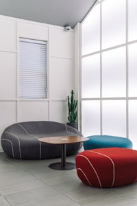 A modern interior space featuring stylish, minimalist furniture including a curved gray sofa, a cylindrical red pouf, and a teal pouf. A round wooden coffee table sits in the center. A large frosted glass wall allows diffused light to fill the room, creating a calm and serene ambiance. A tall green cactus in a pot adds a natural element to the setting.