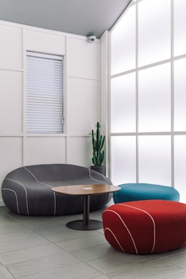 A modern interior space featuring stylish, minimalist furniture including a curved gray sofa, a cylindrical red pouf, and a teal pouf. A round wooden coffee table sits in the center. A large frosted glass wall allows diffused light to fill the room, creating a calm and serene ambiance. A tall green cactus in a pot adds a natural element to the setting.