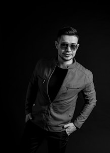 A man wearing sunglasses and a leather jacket stands confidently, leaning slightly to one side against a dark backdrop. His hands are in the pockets of his jacket, and he has a relaxed, self-assured expression.
