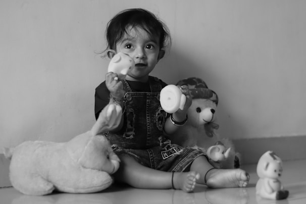 A young child sits against a plain wall, playing with various stuffed animals and toys. The child is wearing denim overalls and has a curious expression while holding a toy. The scene is tranquil and intimate, with soft lighting highlighting the child's features.