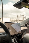 A modern car interior showcasing a large central touchscreen display with a navigation interface. The screen displays a map and vehicle settings. The dashboard is sleek and minimalist, with a prominent steering wheel in the foreground. The background through the windshield shows an industrial area with power lines and structures.