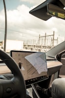 A modern car interior showcasing a large central touchscreen display with a navigation interface. The screen displays a map and vehicle settings. The dashboard is sleek and minimalist, with a prominent steering wheel in the foreground. The background through the windshield shows an industrial area with power lines and structures.