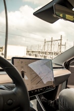 A modern car interior showcasing a large central touchscreen display with a navigation interface. The screen displays a map and vehicle settings. The dashboard is sleek and minimalist, with a prominent steering wheel in the foreground. The background through the windshield shows an industrial area with power lines and structures.