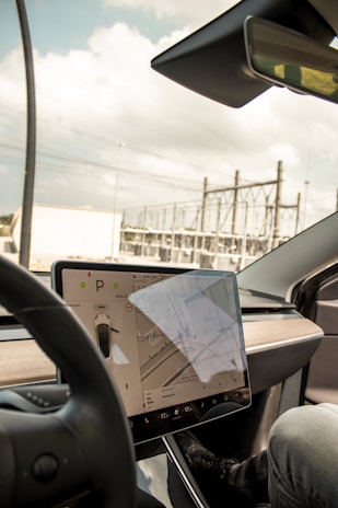A modern car interior showcasing a large central touchscreen display with a navigation interface. The screen displays a map and vehicle settings. The dashboard is sleek and minimalist, with a prominent steering wheel in the foreground. The background through the windshield shows an industrial area with power lines and structures.