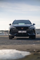 A modern, sleek black car is parked on a road, with a cloudy sky in the background. The vehicle's front is prominently displayed, showcasing its bold and angular design, featuring sharp headlights and a distinctive grille with a prominent logo.