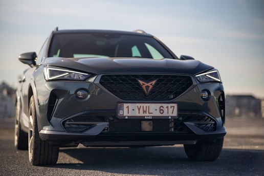 A sleek, modern SUV is parked on a flat surface. The vehicle's front features sharp, angular headlights and a prominent grille with a unique emblem. The license plate is visible, indicating it is from Belgium. The background is slightly blurred, suggesting an urban or open-space setting under a clear sky.