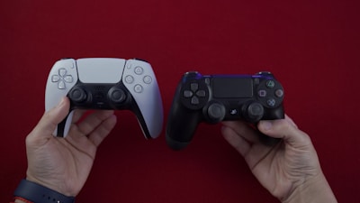 Two hands hold video game controllers against a red background. One controller is a white PlayStation 5 DualSense, and the other is a black PlayStation 4 DualShock.