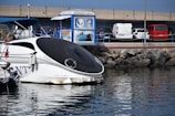 A modern boat with a sleek, black and white design is docked near a marina. Several cars are parked nearby, with a red car and a white van visible. There is a blue information kiosk displaying images of dolphins and marine life. The backdrop includes a stone breakwater and a concrete overpass.