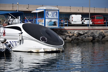 A modern boat with a sleek, black and white design is docked near a marina. Several cars are parked nearby, with a red car and a white van visible. There is a blue information kiosk displaying images of dolphins and marine life. The backdrop includes a stone breakwater and a concrete overpass.