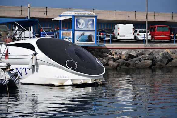 A modern boat with a sleek, black and white design is docked near a marina. Several cars are parked nearby, with a red car and a white van visible. There is a blue information kiosk displaying images of dolphins and marine life. The backdrop includes a stone breakwater and a concrete overpass.