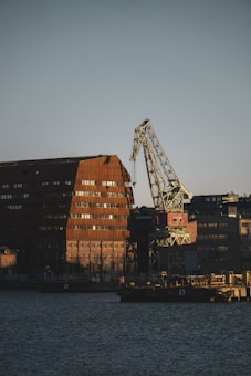 There is an industrial waterfront scene featuring a large crane next to rust-colored building structures. The buildings have a utilitarian design characterized by many small windows. The crane has an angular metal frame and is positioned near the edge of the water. The area appears to be a port or dock with additional smaller buildings and infrastructure visible.