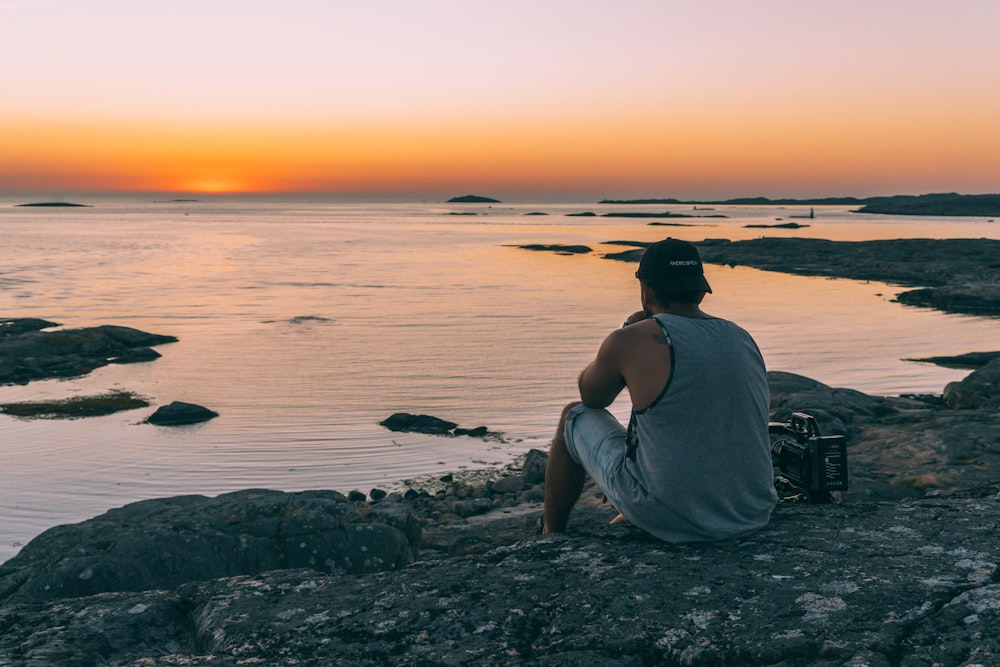 man in gray tank top sitting on rocky shore during sunset