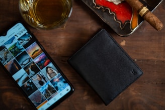 A wooden surface holds a glass of amber liquid, likely whiskey, next to a smartphone displaying a social media gallery filled with photographs. A black leather wallet rests nearby, and there is an ashtray containing an unlit brown cigar.