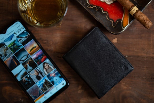 A wooden surface holds a glass of amber liquid, likely whiskey, next to a smartphone displaying a social media gallery filled with photographs. A black leather wallet rests nearby, and there is an ashtray containing an unlit brown cigar.