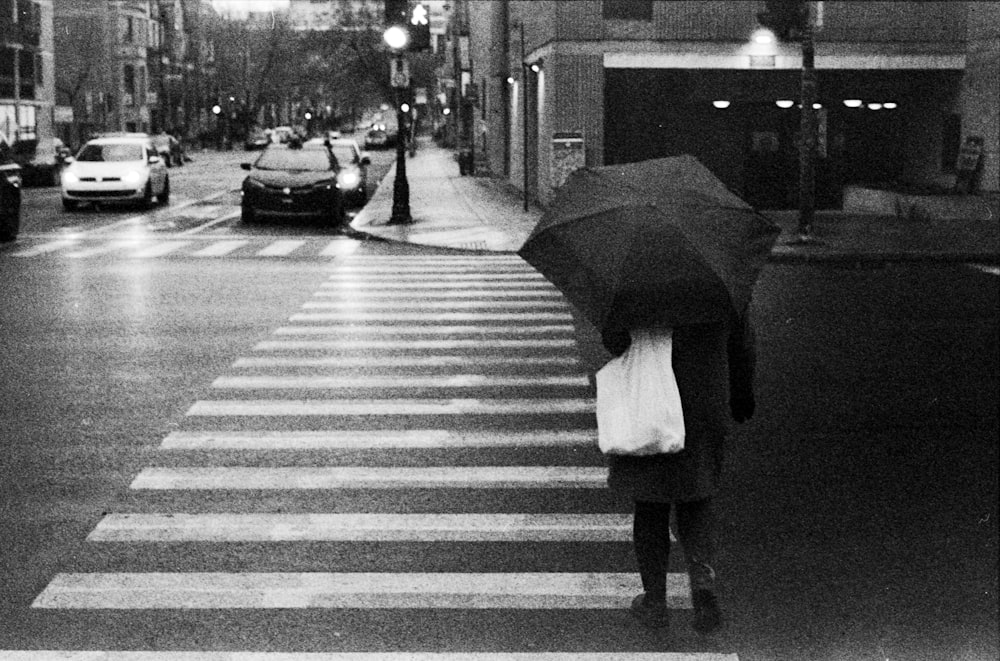 grayscale photo of woman in white dress holding umbrella walking on pedestrian line