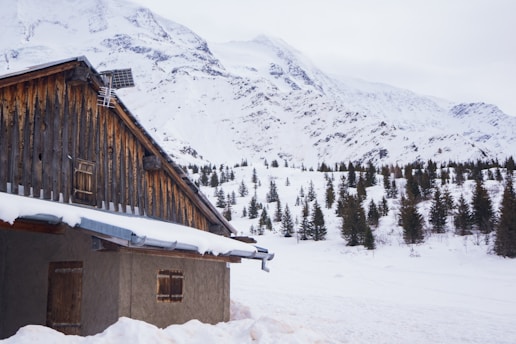 A rustic wooden cabin is nestled in a snowy landscape with a mountain range in the background. Pine trees dot the landscape, and the cabin's roof is covered with snow. A solar panel is mounted on the roof of the cabin.