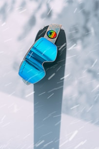 A pair of vibrant blue ski goggles is perched atop a snowboard standing upright against a snowy background. Snowflakes are actively falling, creating a dynamic and wintry scene.