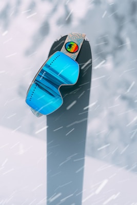 A pair of vibrant blue ski goggles is perched atop a snowboard standing upright against a snowy background. Snowflakes are actively falling, creating a dynamic and wintry scene.