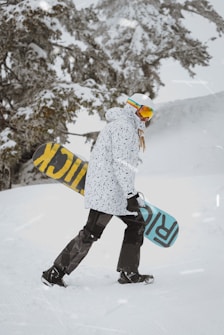 A person dressed in winter sports gear, including a speckled white jacket, dark pants, and white helmet, is carrying a snowboard. The snowboard has large text with contrasting colors on each side. They are walking on a snow-covered landscape with snow-laden trees in the background.
