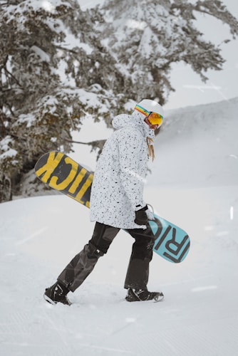 A person dressed in winter sports gear, including a speckled white jacket, dark pants, and white helmet, is carrying a snowboard. The snowboard has large text with contrasting colors on each side. They are walking on a snow-covered landscape with snow-laden trees in the background.