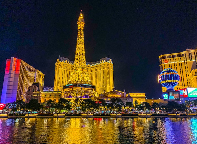 A vibrant nighttime cityscape featuring a brightly illuminated replica of the Eiffel Tower surrounded by colorful lights and buildings. The reflection of these lights shimmers on the water in the foreground, creating a lively and picturesque scene.