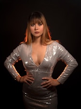 A woman with long hair and bangs stands confidently with her hands on her hips. She is wearing a shimmering silver dress with long sleeves, set against a dark background, creating a striking contrast.