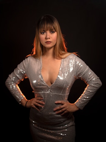A woman with long hair and bangs stands confidently with her hands on her hips. She is wearing a shimmering silver dress with long sleeves, set against a dark background, creating a striking contrast.